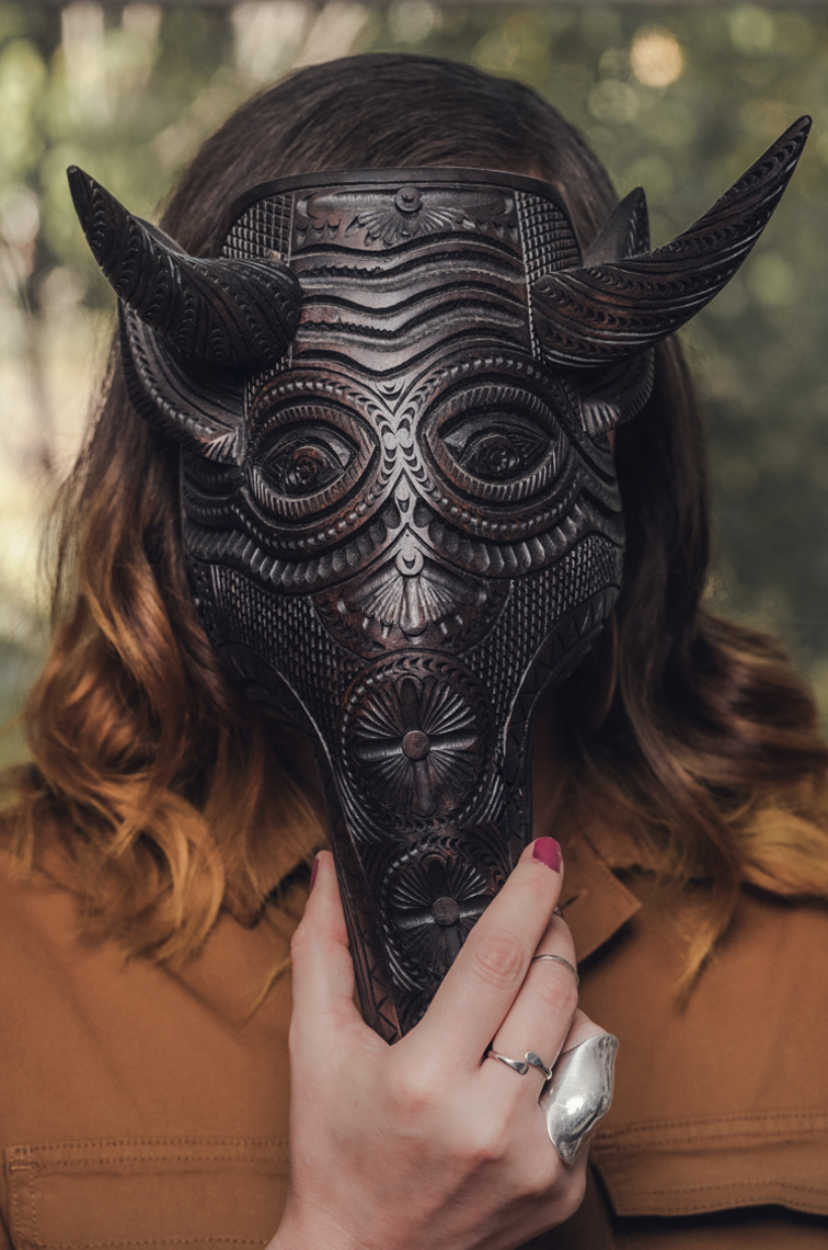 Christine holding a wooden mask of an armadillo with horns that covers her face. The wooden mask was made by a Mexican artist known to make masks for the annual Día de Muertos celebration.